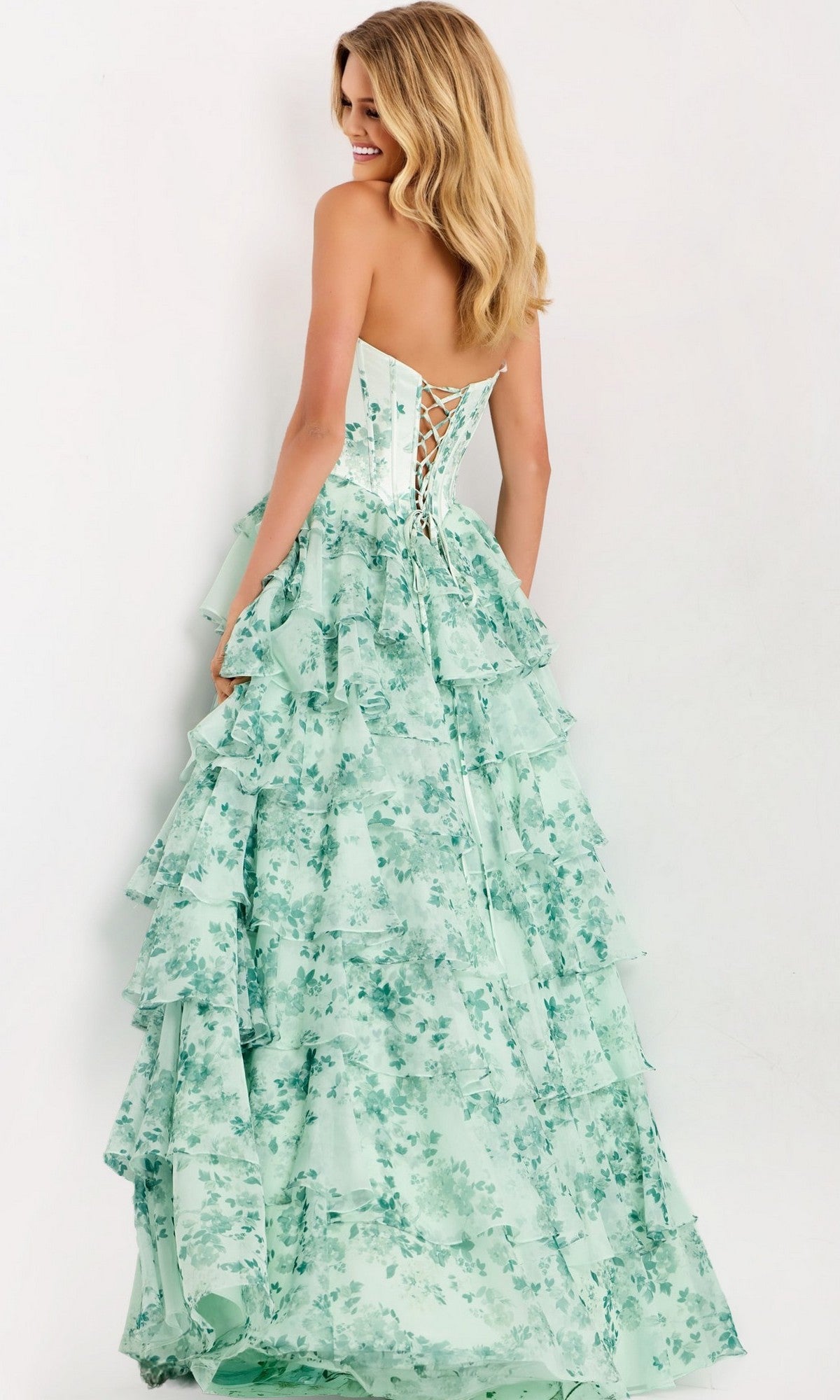 A woman wears the Jovani 48772 long prom dress, a mint green strapless gown with floral print, corset bodice back, and tiered ruffles, posed against a plain background.